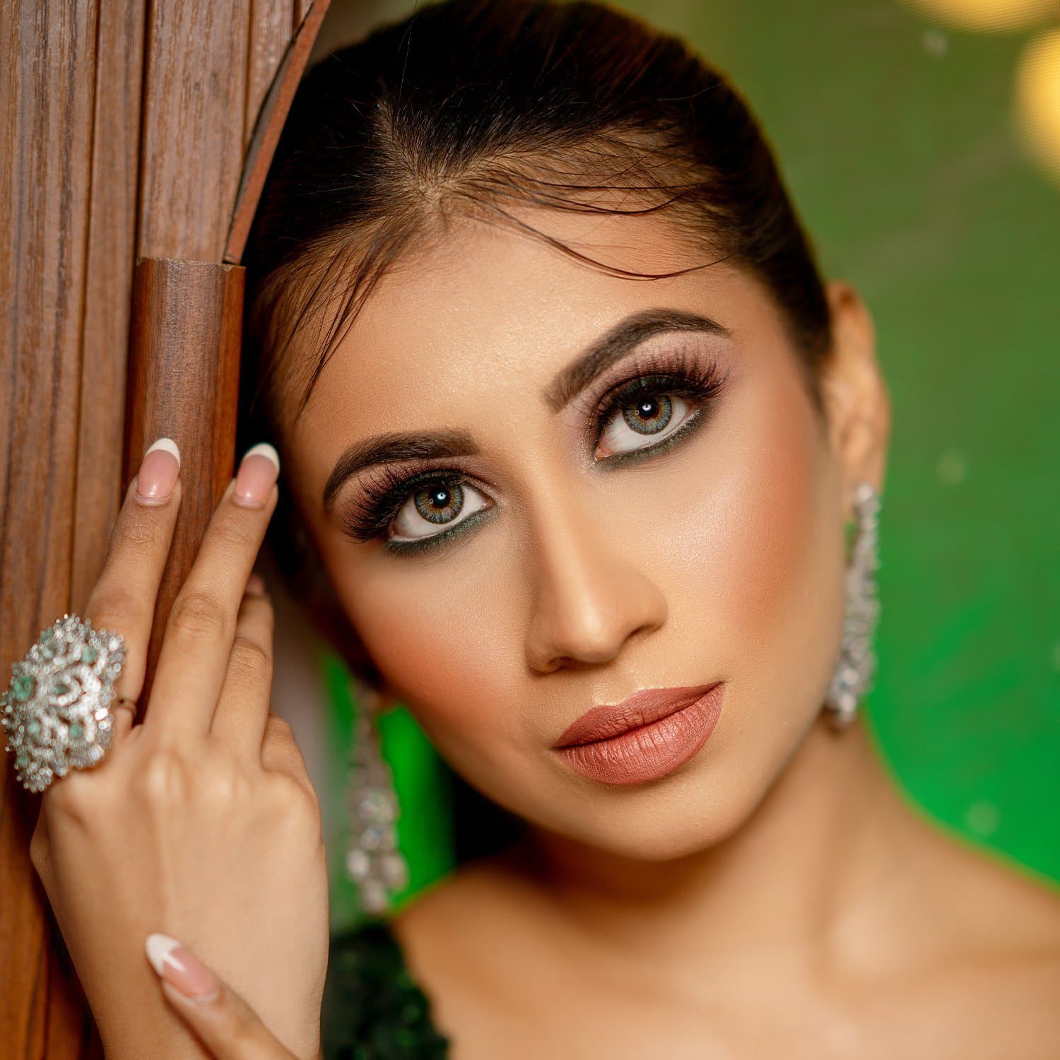 Glam portrait with defined brows, smoky lashes and a nude lip against green drapes